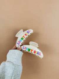 Colorful hair clip with 'LUCKY GIRL' text held against a beige background