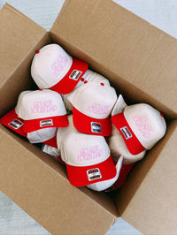 A box full of beige trucker hats with red bills that have 'Go Ask Your Dad' embroidered in pink thread.