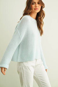 Woman wearing a light blue knitted sweater and white pants against a plain background