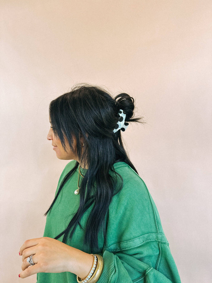 Woman with long black hair styled in a side braid with a decorative hair clip, wearing a green jacket against a beige background.