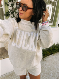 Person wearing an ash sweatshirt with 'LUCKY' printed on it, standing outdoors.