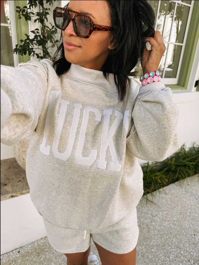 Person wearing an ash sweatshirt with 'LUCKY' printed on it, standing outdoors.