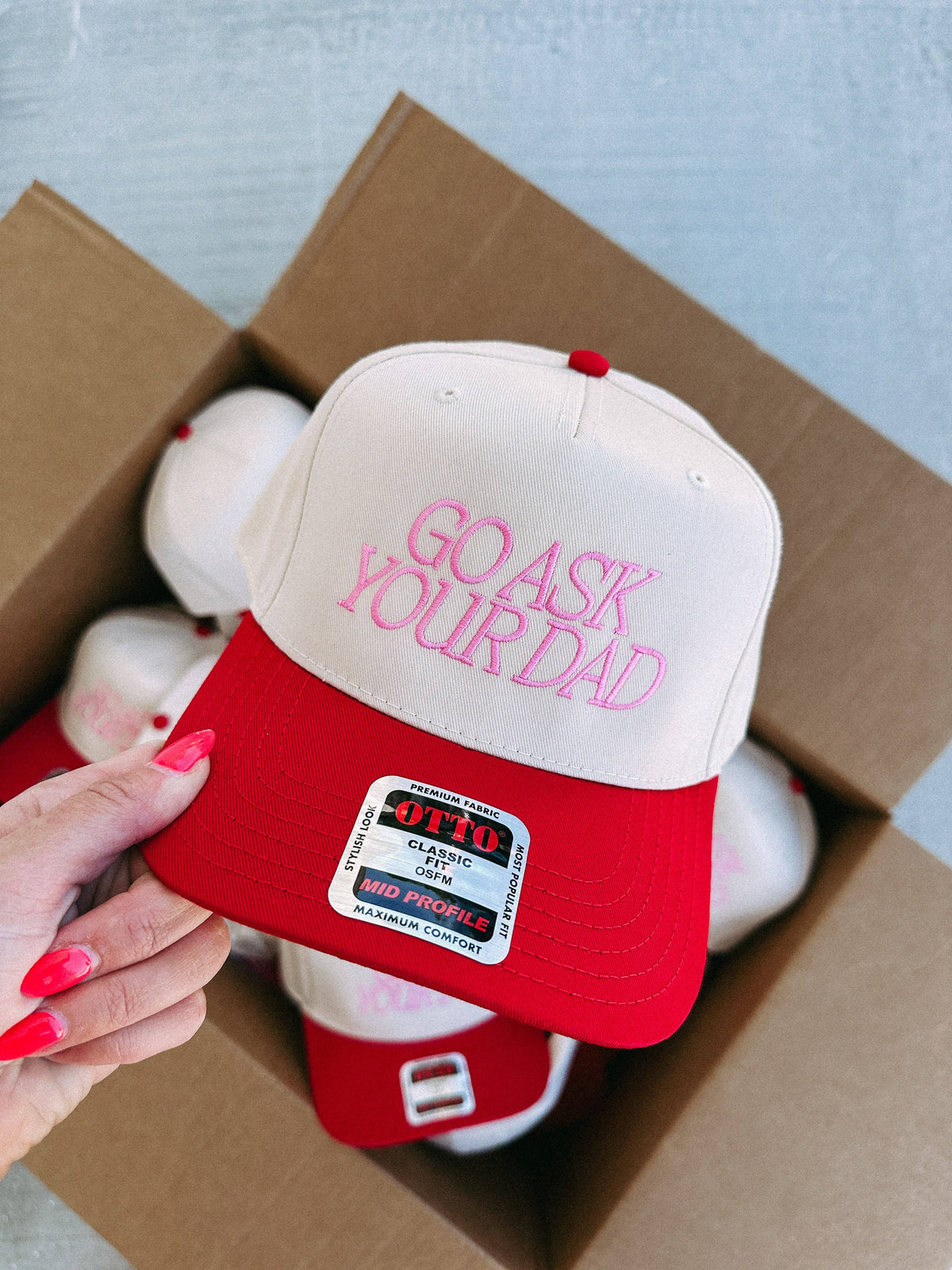 Red bill on a beige trucker hat with 'Go Ask Your Dad' embroidery in pink thread, held by a hand. A box with more caps in the background.